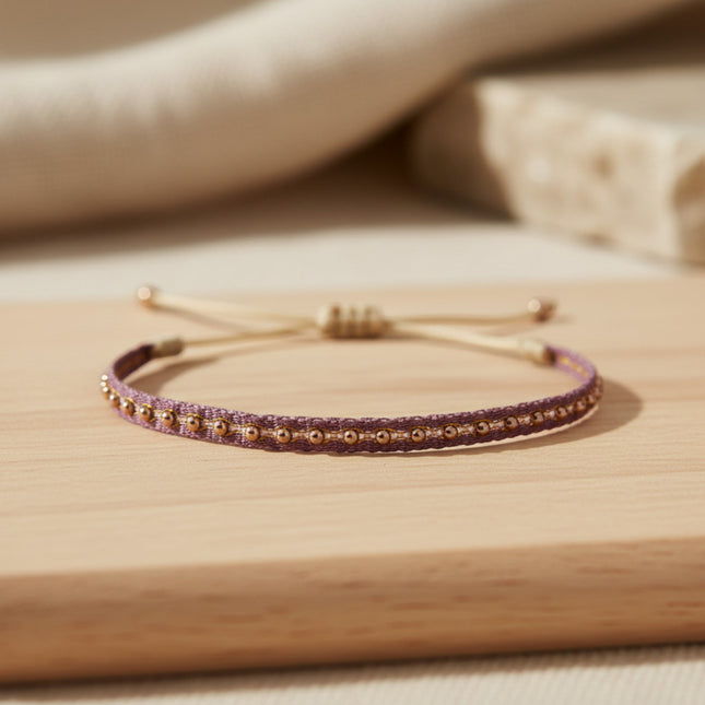 Bracelet fin tissé main Horizons Boutique en tons violets avec billes dorées, cordon ajustable, style bohème chic.