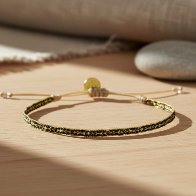 Bracelet fin tissé main Guanabana en tons or et noir, ajustable, avec cordon ciré et breloque dorée.